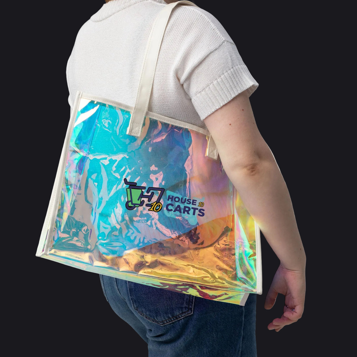 A person wearing a white t-shirt and blue jeans carries a large, semi-transparent holographic tote bag with cream-colored shoulder straps. The bag features a colorful iridescent print with a 'House of Carts' logo printed on the front.