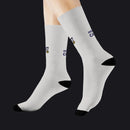 A pair of crew socks worn on a person's feet, shown against a plain white background. The socks are light gray with black heels, toes, and heels, and feature a small navy and gold logo with the text 'HOT CAKE' on the upper calf.