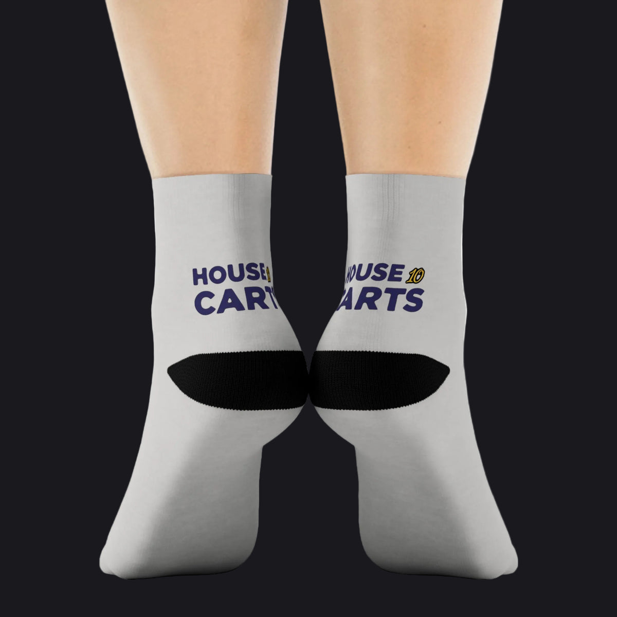 A pair of light gray crew socks worn on feet, featuring a black arch support band and the text 'HOUSE OF CARTS' in navy blue and yellow on the sides.