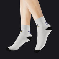 A pair of white crew socks worn on a model's feet, featuring black toe and heel caps, a small gold crest, and navy blue text that reads 'HOT CAKE' on the sides.