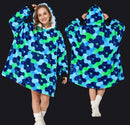 A woman models a hooded lounge blanket/pod in a studio setting, shown from the front and back, with inset close-ups of the hood and front pocket area. The product has a bold blue and green abstract pattern with a white sherpa-like hood lining and black cuffs.